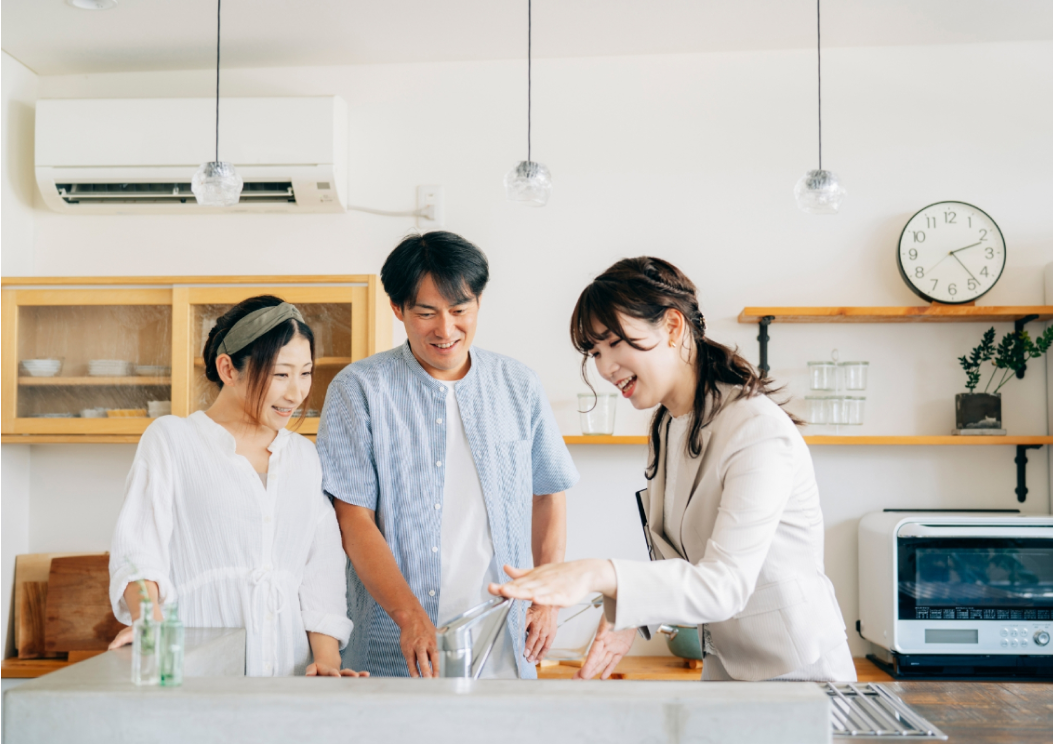 キッチンで蛇口を操作しながら説明する女性と、それを笑顔で見守る夫婦。
