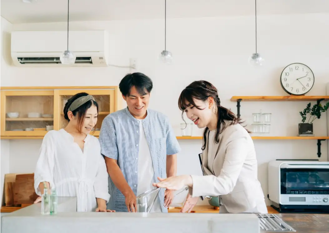 キッチンで蛇口を操作しながら説明する女性と、それを笑顔で見守る夫婦。