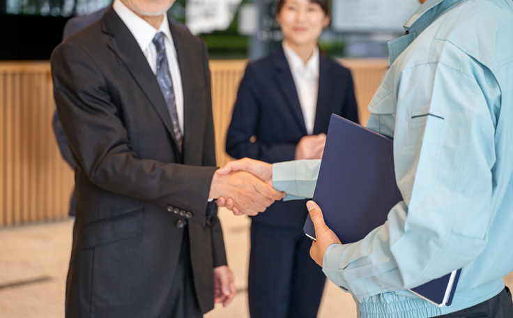 ビジネスシーンでスーツ姿の人物と作業服姿の人物が握手をしている様子。